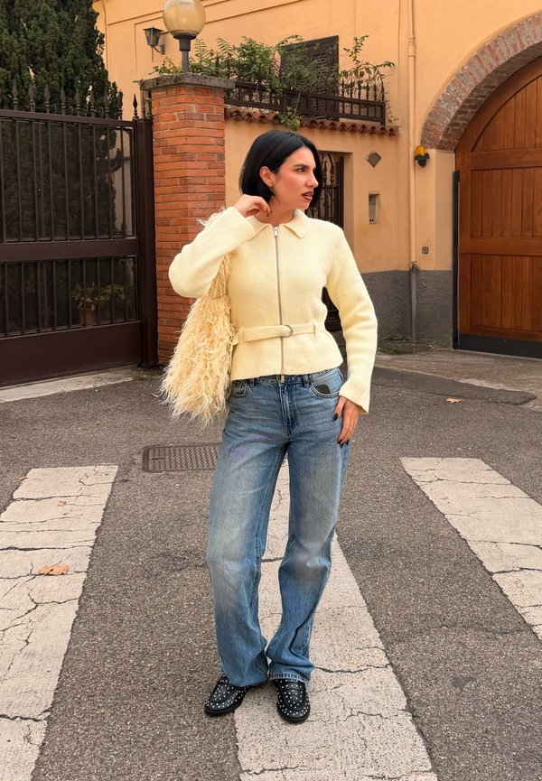 Cream-coloured zip-up jacket with a collar and belt, teamed with light blue wide-leg jeans and black studded loafers. Complemented by a textured bag.