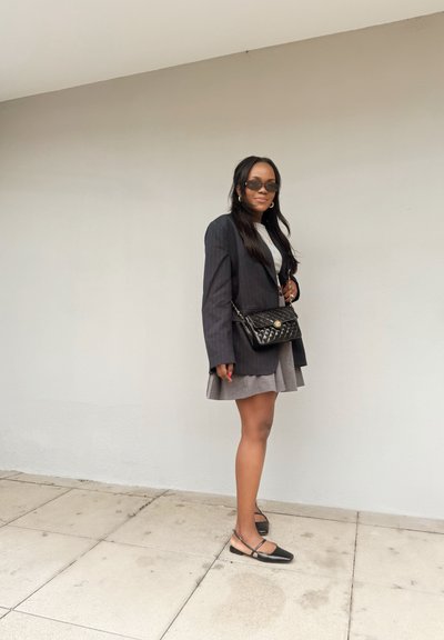 Blazer negro de rayas sobre un vestido gris claro, bolso negro acolchado, bailarinas negras con tiras al tobillo y cabello largo y oscuro. Fondo de hormigón.