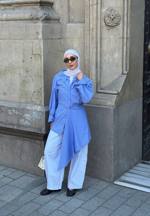 Kvinde iført hvid hijab, blåstribet asymmetrisk skjorte, hvide bukser, sorte sko og solbriller, der står ved hjørnet af en stenkonstruktion på en brolagt gade.