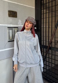 Svetlošedá bunda na zips so bielymi pruhmi na rukávoch, logo Adidas na hrudi, skombinovaná so �šedými teplákmi. Kockovaná čiapka a náušnice na kruhu.