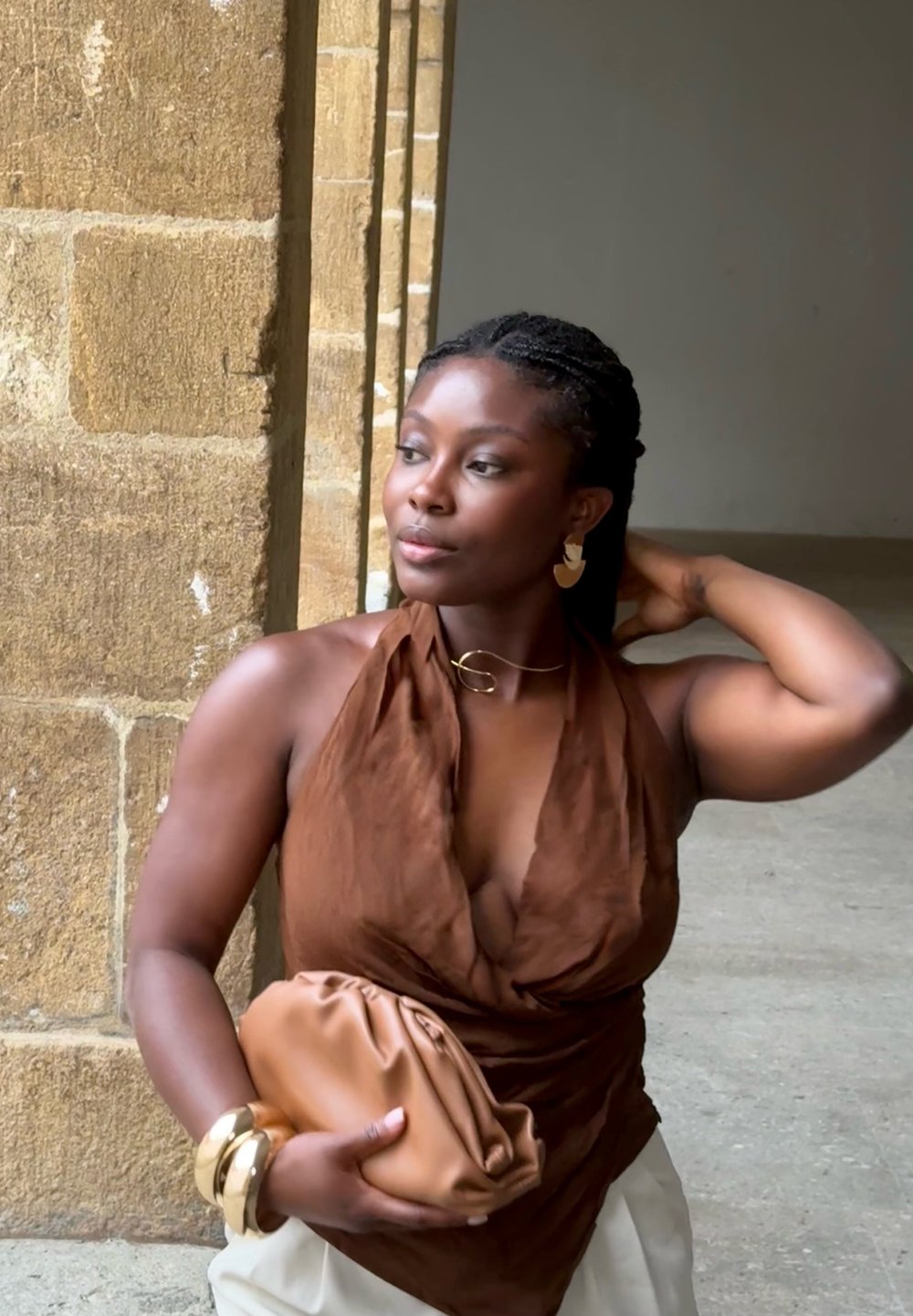 Haut halter en drapé marron, bijoux en or, pochette beige et bracelets empilés. Peau lisse et cheveux tressés contre un mur en pierre texturé.