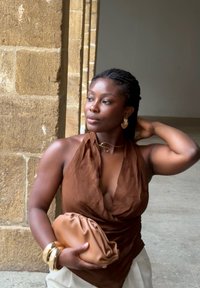 Haut halter en drapé marron, bijoux en or, pochette beige et bracelets empilés. Peau lisse et cheveux tressés contre un mur en pierre texturé.