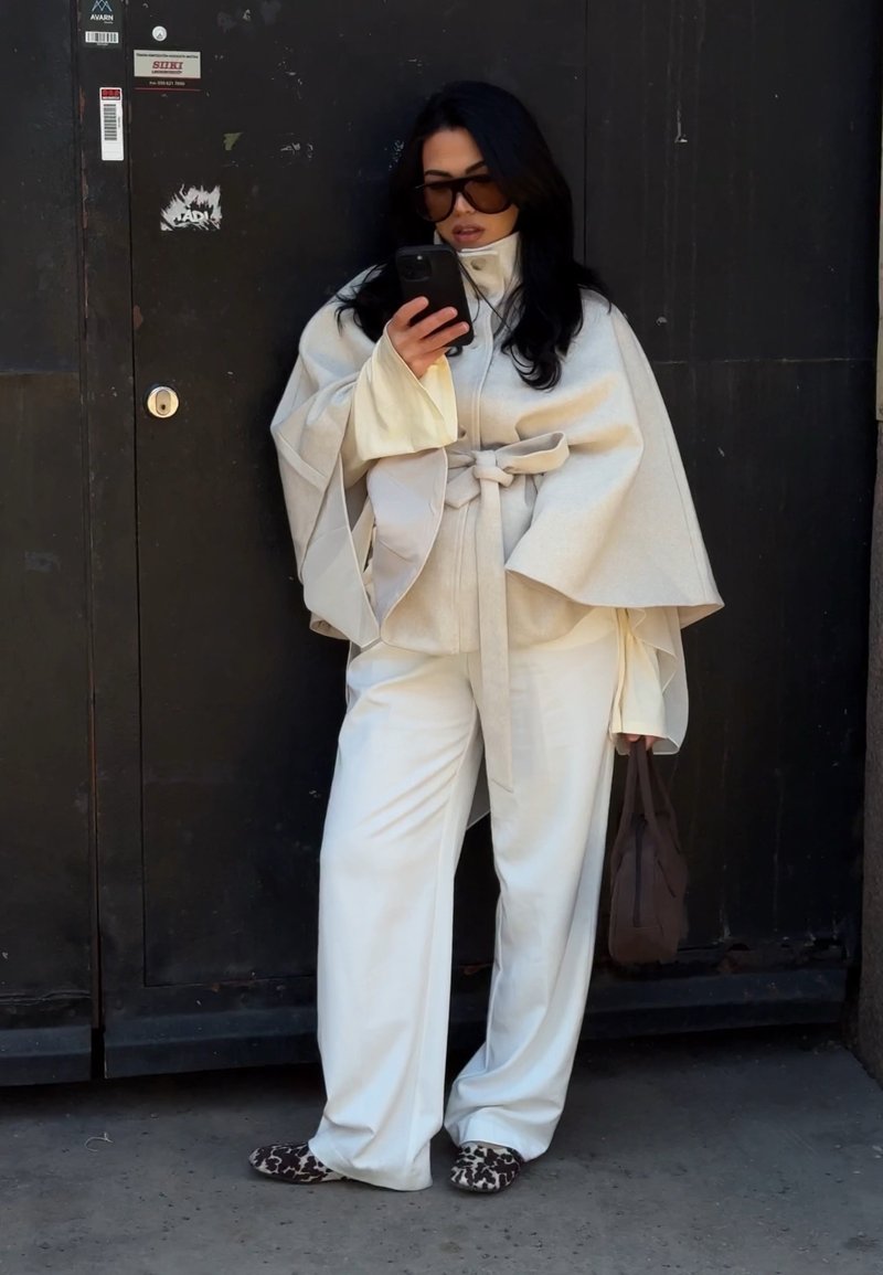 Femme aux longs cheveux bruns et lunettes de soleil, portant une cape beige, un pantalon blanc ample, des chaussures à imprimé léopard, tenant un téléphone et un sac marron, debout près d'une porte noire.