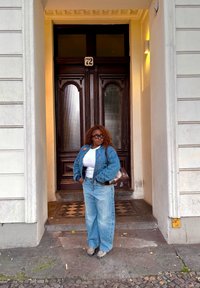 En denimjakke over en hvit skjorte, lyseblå baggy jeans og mønstrede sko; står foran doble tre dører med nummer 72.