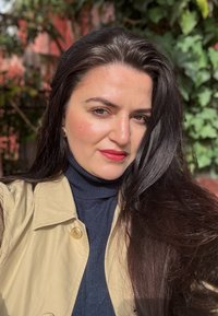 Beige trenchcoat med gullfester over en mørkeblå genser med høy hals. Langt, mørkt hår. Naturlig omgivelser med grønt i bakgrunnen.