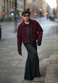 Giacca testurizzata bordeaux con zip, t-shirt nera e pantaloni a righe sagomati. Guanti neri senza dita. Sfondo urbano.