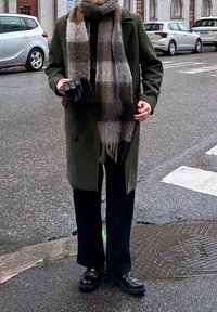 Manteau vert olive, écharpe à motifs gris et brun, gants noirs, pantalon foncé et chaussures noires. Rue mouillée et voitures stationnées en arrière-plan.