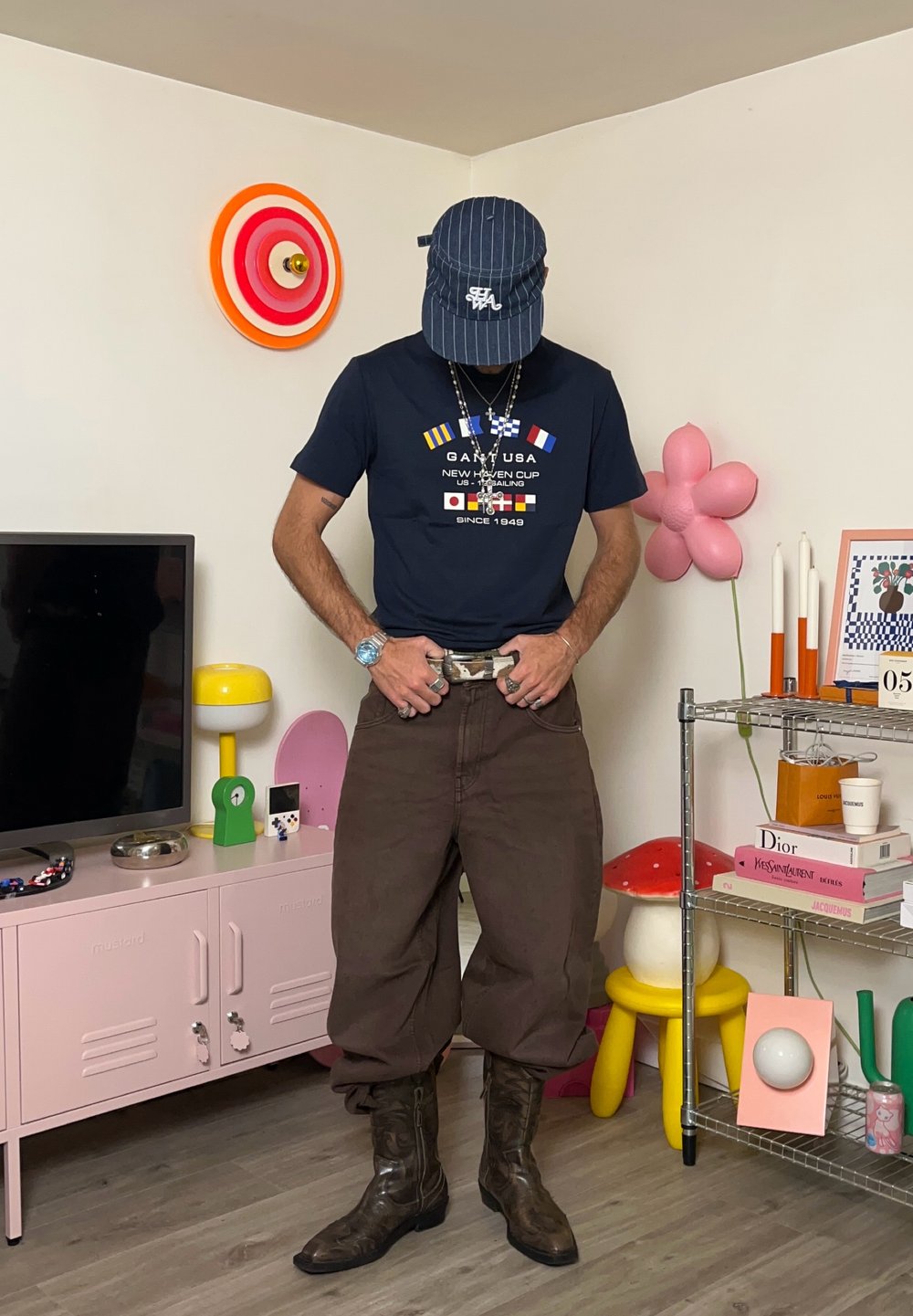 Marineblå t-shirt med grafisk print, brune brede bukser, brune cowboy støvler. Accessories inkluderer halskæder og en blåstribet kasket.