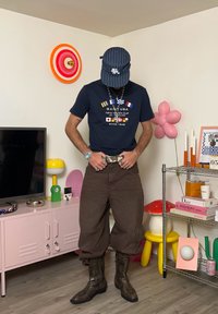 Marineblå t-shirt med grafisk print, brune brede bukser, brune cowboy støvler. Accessories inkluderer halskæder og en blåstribet kasket.