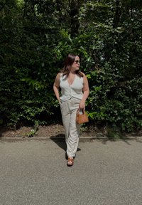 Haut sans manches gris clair avec des boutons, pantalon large crème, sac à main marron et sandales marron, sur fond de feuillage verdoyant.