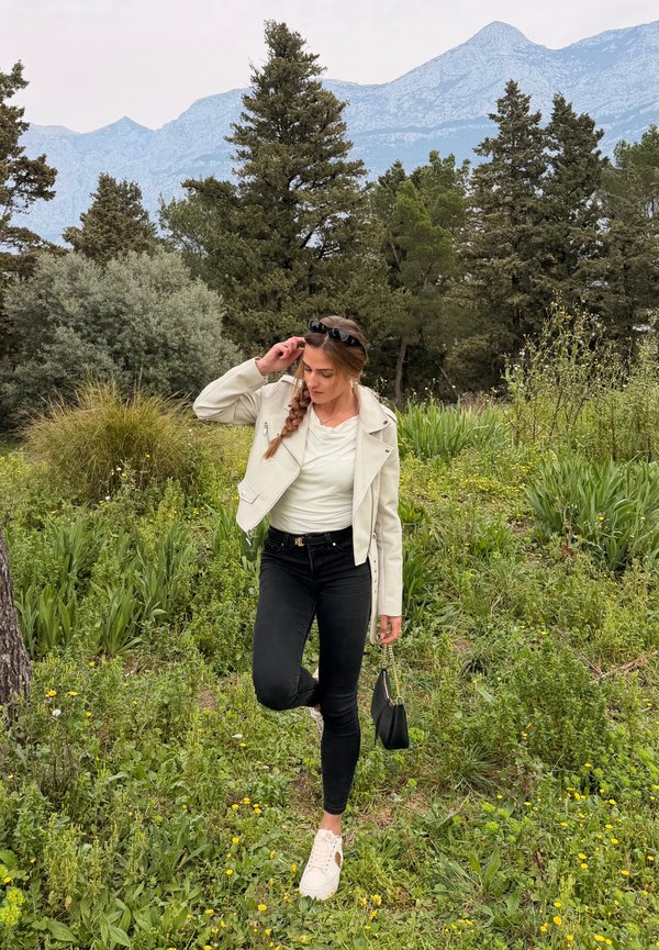 Kvinde i hvid jakke, sorte bukser og hvide sneakers står på ét ben i et græsareal med træer og bjerge i baggrunden.