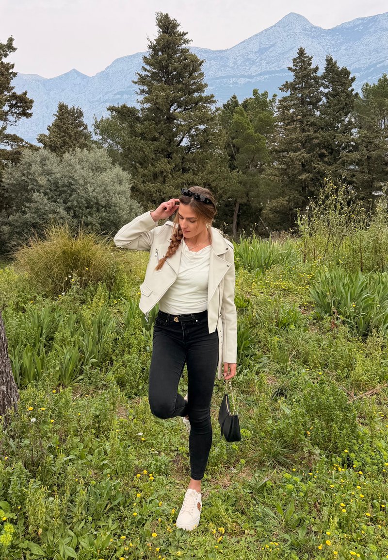 Nainen valkoisessa takissa, mustissa housuissa ja valkoisissa tennareissa seisoo yhdellä jalalla ruohoisella alueella, taustalla puita ja vuoria.