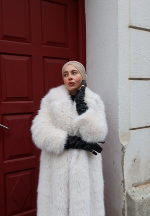 Mujer con pañuelo beige en la cabeza y abrigo de piel blanco, posando con guantes de cuero negros junto a una puerta roja y una pared blanca texturizada.