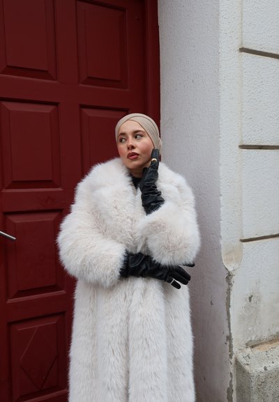 Mujer con pañuelo beige en la cabeza y abrigo de piel blanco, posando con guantes de cuero negros junto a una puerta roja y una pared blanca texturizada.