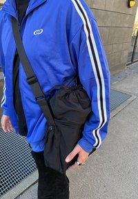 Μαύρη τσάντα με κορδόνι κρατημένη κάτω από το χέρι; άνδρας με μπλε σακάκι με λευκές και μαύρες ρίγες στα μανίκια; υφασμάτινη υφή; αστικό περιβάλλον.