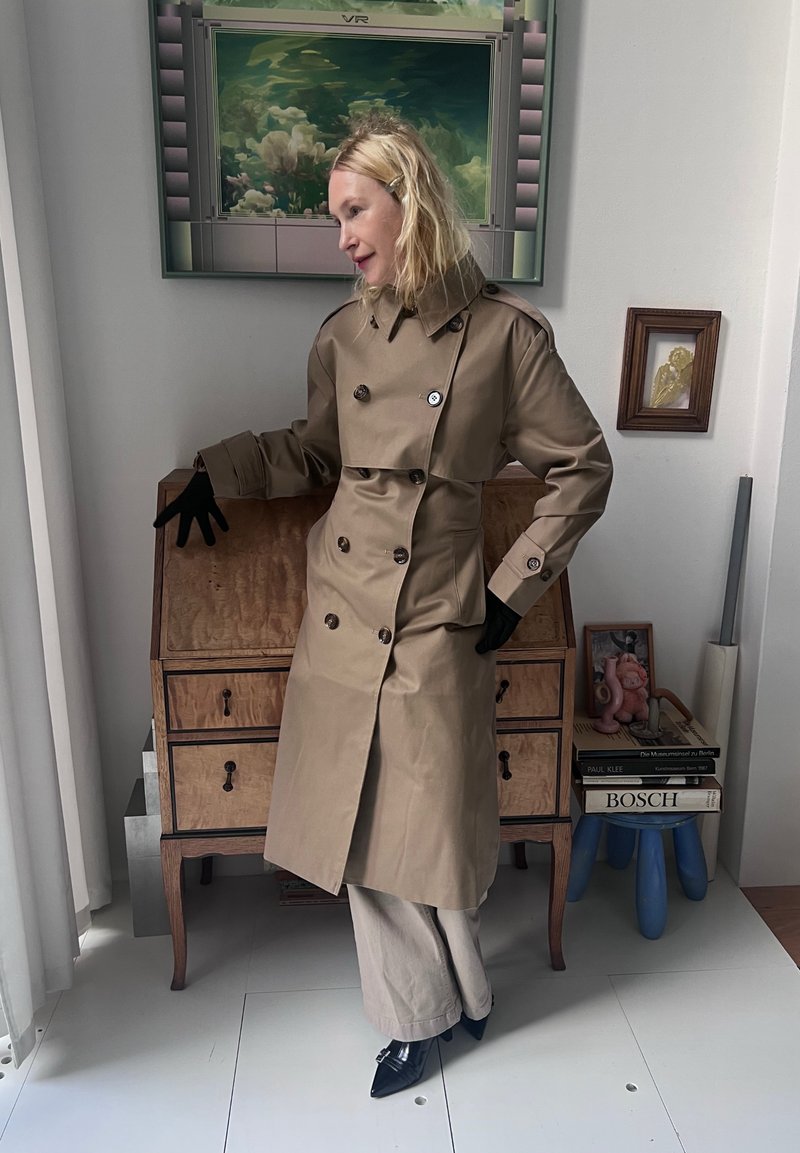 Beige Trenchcoat mit doppelreihigen Knöpfen, hohem Kragen und breiten Manschetten. Getragen mit schwarzen Handschuhen, hellen Hosen und spitzen schwarzen Schuhen.
