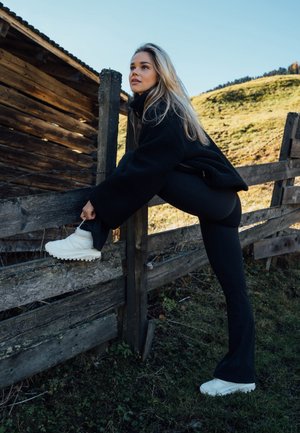 Botas blancas gruesas hasta el tobillo con superficie texturizada y suelas gruesas, combinadas con leggings negros acampanados y una chaqueta de forro polar negra, frente a una cerca de madera.