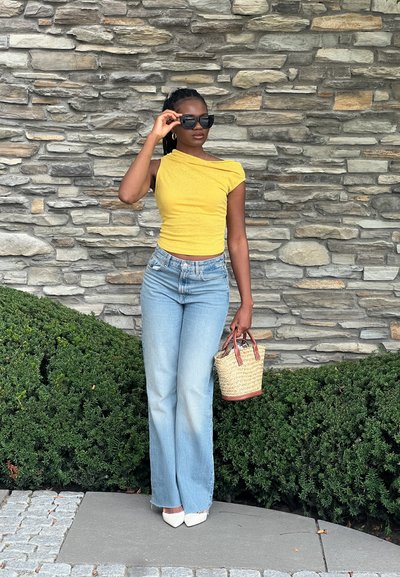 Top amarillo sin hombros, jeans acampanados de color azul claro, tacones puntiagudos blancos, sosteniendo un bolso de fibra con acentos rojos, frente a un fondo de pared de piedra.