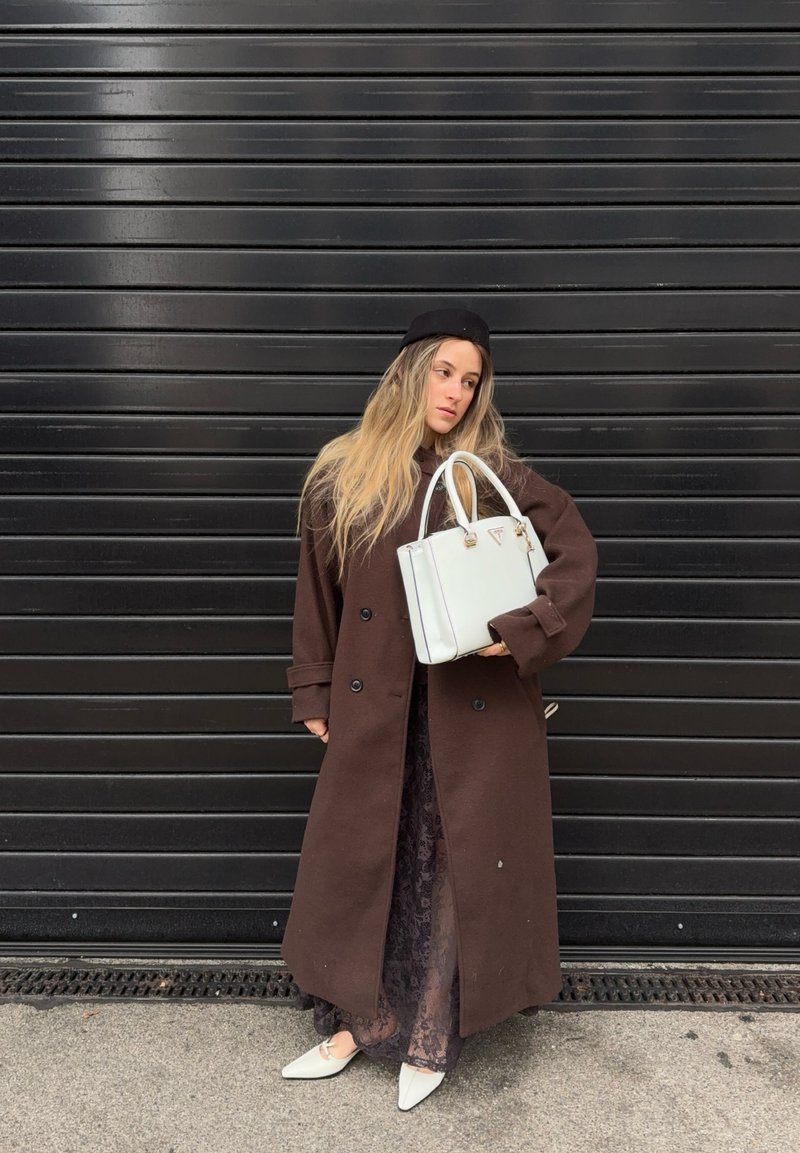 Kvinde iført lang brun frakke og sort baret, der holder en hvid håndtaske og står foran en sort bølgepladet metalport.