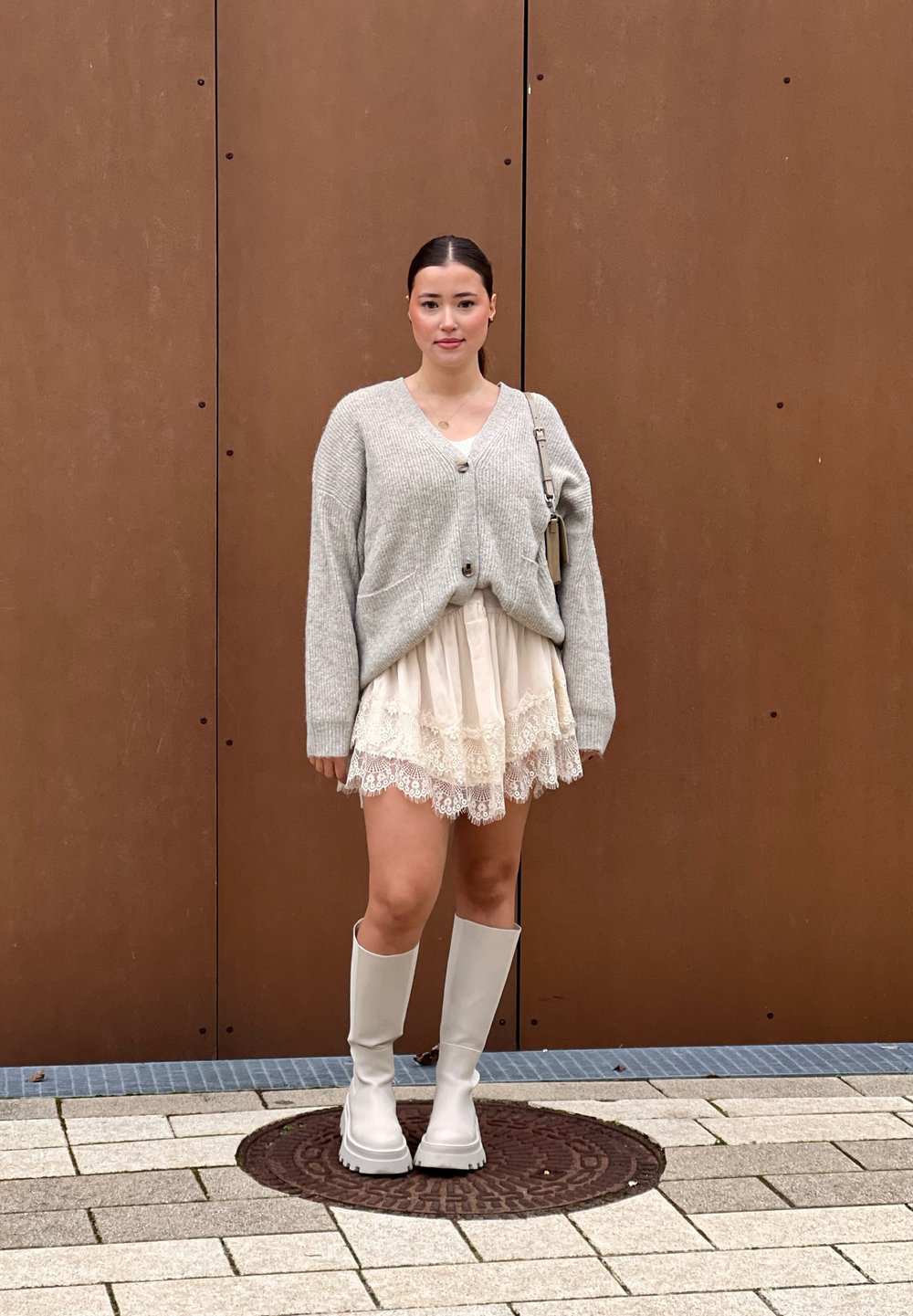 Jasnoszary kardigan z guzikami, nałożony na spódnicę w kolorze kremowym ozdobioną koronką. Całość dopełniają białe kozakami do kolan. Gładkie brązowe tło.