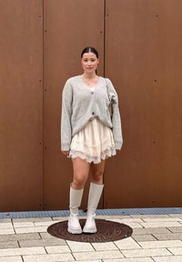 Jasnoszary kardigan z guzikami, nałożony na spódnicę w kolorze kremowym ozdobioną koronką. Całość dopełniają białe kozakami do kolan. Gładkie brązowe tło.