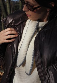 Giacca imbottita marrone con finitura lucida, chiusura frontale con zip; maglione lavorato a maglia color crema con rifiniture grigie e trama. La mano di una persona tocca la giacca.