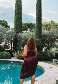Burgunderfarbendes Satin-Kleid mit enganliegender Silhouette, knielang und dünnen Trägern, am Poolrand inmitten von Grün und Steindekor präsentiert.