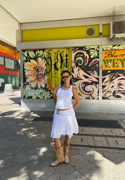 Top blanco sin mangas y falda, botas de vaquero marrones, con un hebilla de cinturón circular. Fondo de graffiti colorido que presenta flores y diseños abstractos.