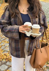 Bruine geruite jas met grote mouwen, houdt twee koffiekopjes vast in een beige houder. Bruine tas met een elegante vormgeving. Witte broek zichtbaar.