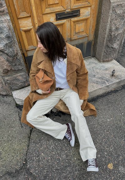 Abrigo de ante marrón, camisa blanca, pantalones crema y zapatillas marrones. Sosteniendo una bolsa color canela. Sentado sobre una superficie de piedra contra una puerta de madera.