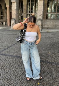 Borsa nera con borchie argentate, top bianco a tubino, jeans azzurri a gamba larga con accenti strappati e sandali neri su pavimentazione in ciottoli.