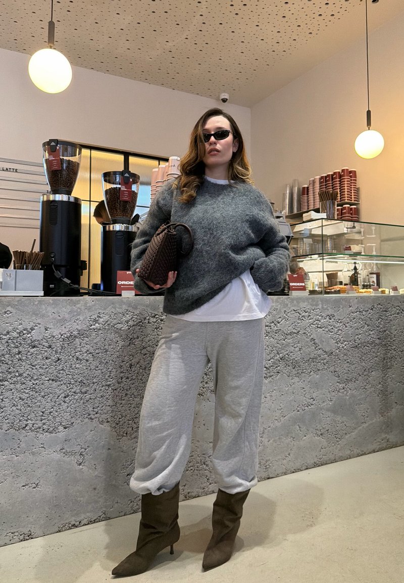 Femme portant des lunettes de soleil, un pull gris, et un pantalon ample tenant un sac, debout dans un café moderne avec des machines à café et une vitrine en arrière-plan.