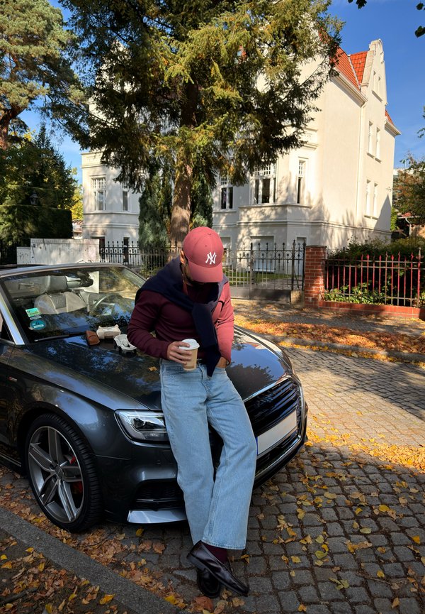 Un hombre con una gorra roja y un jersey burdeos se apoya en un coche negro mientras sostiene un vaso para llevar, en una calle cubierta de hojas.