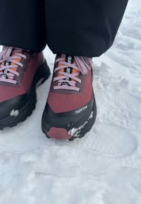 Chaussures de randonnée bordeaux et noires en tissu texturé, avec des lacets violets et un accent orange. De la neige est visible sur les semelles, sur un sol enneigé.