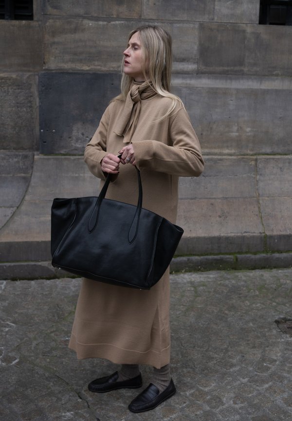 Svart läder tote-väska med förlängda handtag, matchad med en beige, långärmad klänning med en rosett vid halsen. Platta skor och strumpor syns.