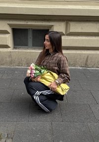 Oseba prikleči na pločniku, oblečena v rjavo karirasto srajco in črne trenirke z belimi črtami. Drži šopek in zlat nahrbtnik.