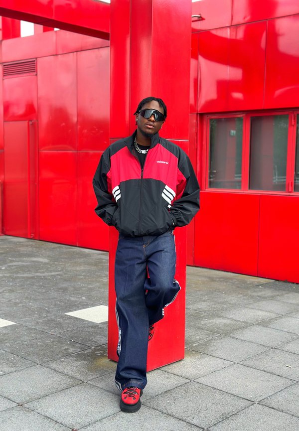 Joven con gafas de sol y chaqueta Adidas rojo-negra apoyado en un pilar rojo brillante en un entorno urbano.