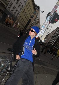 Person iført en blå lue, solbriller og en blå jakke med racingtema, og svarte løstsittende jeans, som står ved siden av en sykkel.