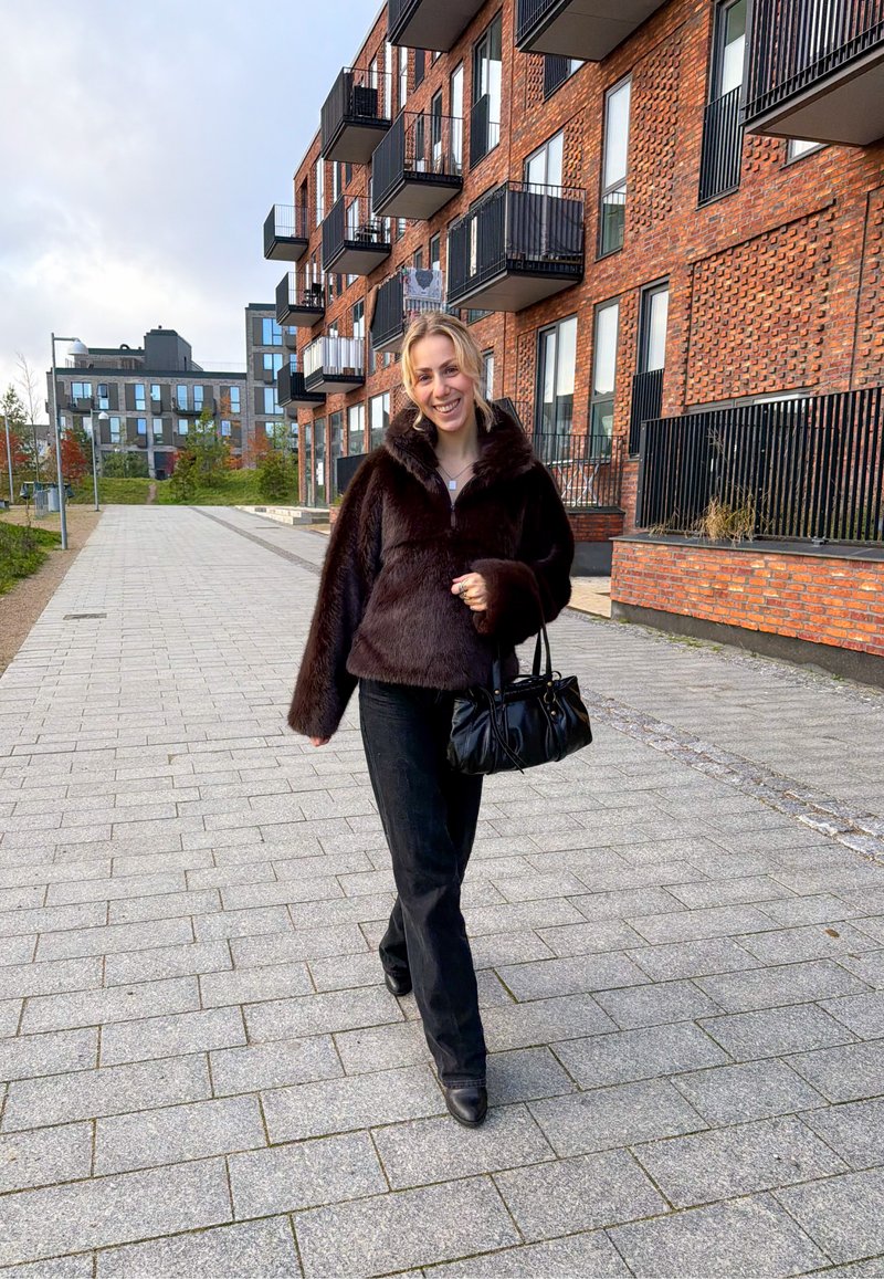 Giacca in pelliccia ecologica marrone con collo alto, jeans neri e borsa nera. Sullo sfondo edifici moderni in mattoni e una passerella lastricata.