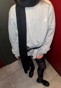 Šedá mikina s dlhým čiernym pleteným šálom, čiernym opaskom, priehľadnými čiernymi pančuchami s čipkovaným lemom a masívnymi čiernymi členkovými čižmami.