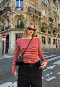 Rot- und weiß gestreiftes Langarmshirt, kombiniert mit schwarzen hochgeschnittenen Hosen. Schwarze Schultertasche und Sonnenbrille. Urbane Straßenszene.