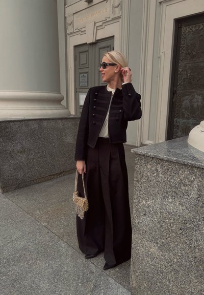 Chaqueta negra recortada con botones decorativos, combinada con pantalones negros de talle alto. Sosteniendo un bolso tejido beige, con arquitectura de mármol de fondo.