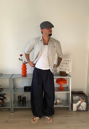 Hombre con gorra gris, camiseta blanca sin mangas, camisa a cuadros, pantalones anchos negros y chanclas, está frente a una estantería con discos y decoración.