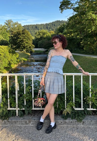 Top sin mangas de volantes azul, mini falda de cuadros gris, mocasines negros, sosteniendo un bolso estampado, de pie cerca de un arroyo con vegetación.