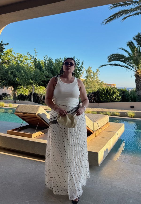 Kvinde iført en hvid tanktop og prikket nederdel står ved en pool med liggestole, omgivet af træer og klar blå himmel.
