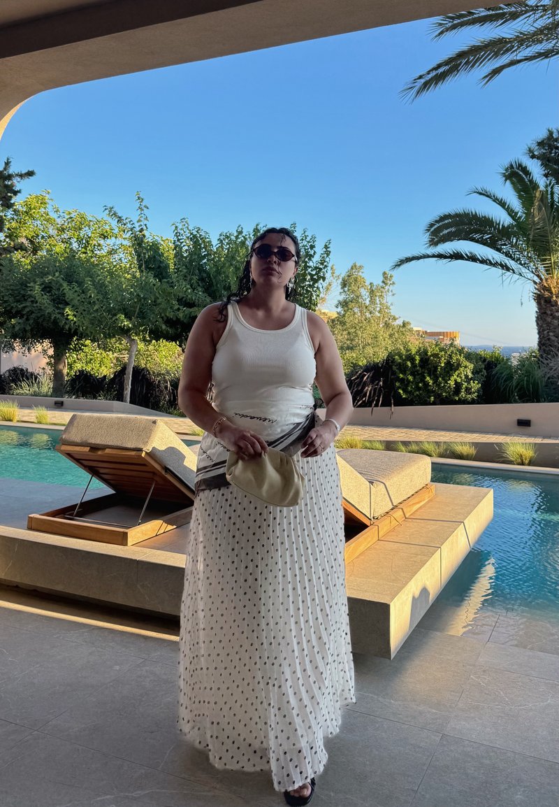 Femme portant un débardeur blanc et une jupe à pois se tient près d'une piscine avec des chaises longues, entourée d'arbres et d'un ciel bleu clair.