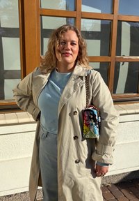 Béžový trenčkot s velkými knoflíky přes světle modré tričko a džíny. Barevná vzorovaná kabelka s řetízkem a korálky.