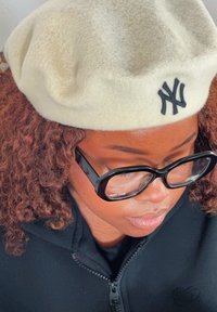 Berretto di lana bianco con logo NY, abbinato a grandi occhiali neri, indossato con una giacca nera con zip. Prospettiva ravvicinata.