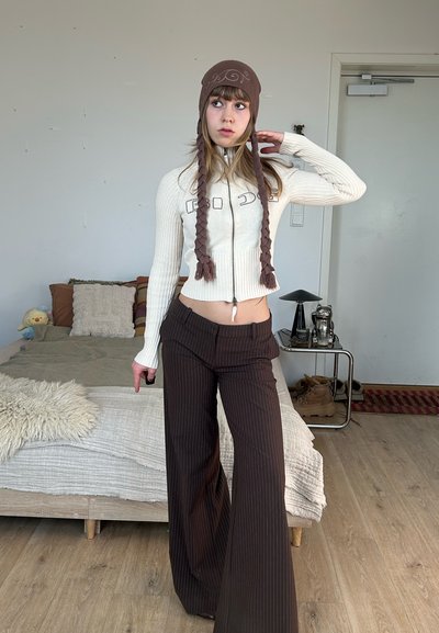 Joven con un sombrero marrón y pantalones de rayas está de pie en un dormitorio con una cama y una mesita de noche al fondo.