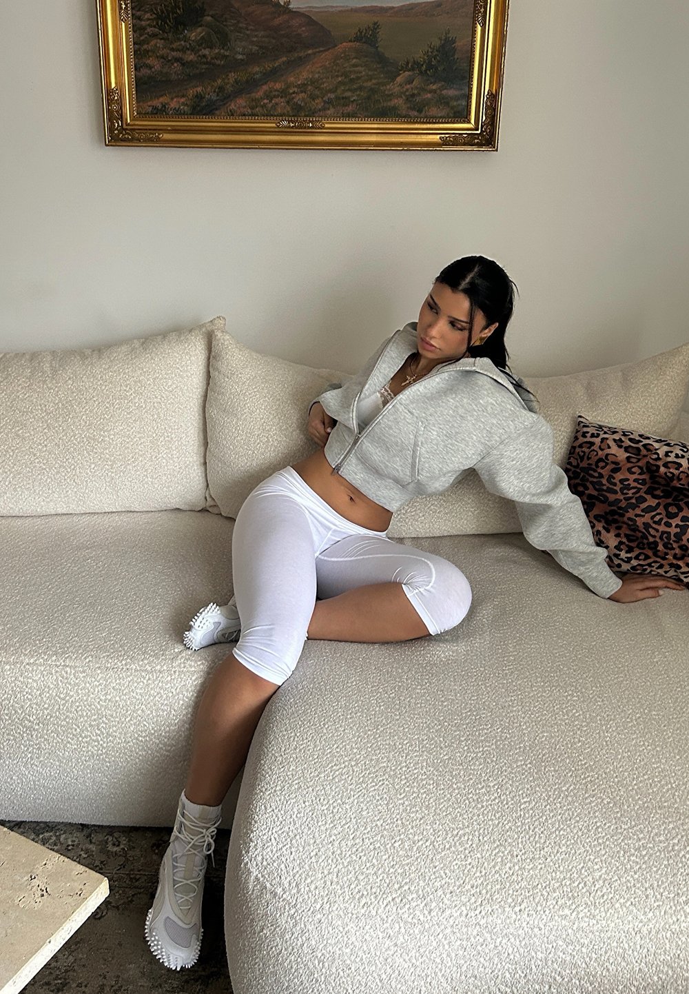 Sweat à capuche gris court, leggings blancs et bottes de couleur claire. Canapé beige clair texturé avec un design confortable. Coussin à imprimé léopard à proximité.